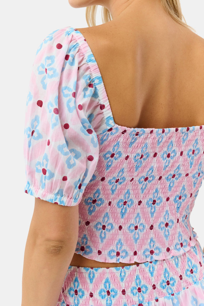 Wearing the Lerato Dovie Top by Roller Rabbit, a woman smiles with her hand on her hip. The light pink cotton crop top has a sweetheart neckline and features a blue and red floral geometric pattern.