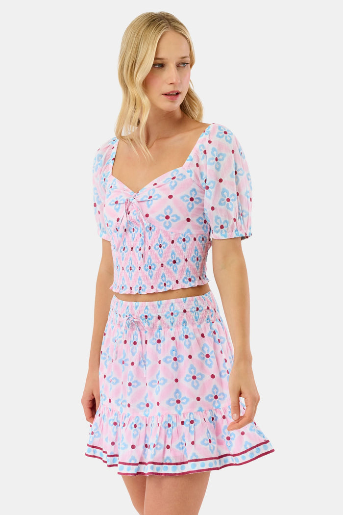 Wearing the Lerato Dovie Top by Roller Rabbit, a woman smiles with her hand on her hip. The light pink cotton crop top has a sweetheart neckline and features a blue and red floral geometric pattern.