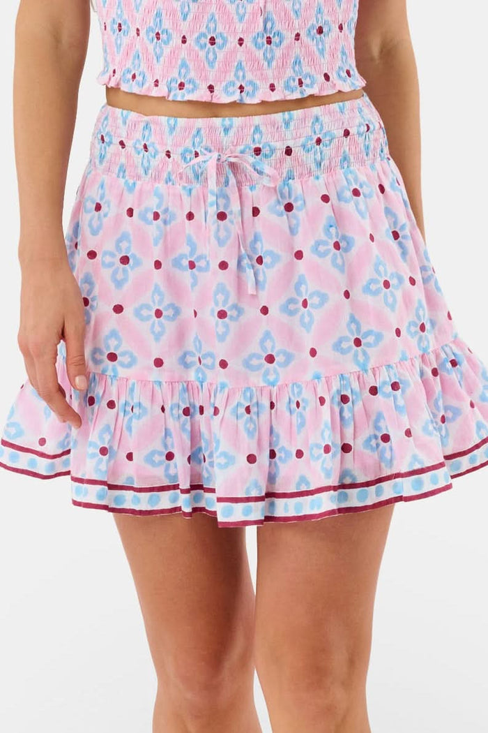 A woman wears the Roller Rabbit Lerato Peyton Skirt, a pink cotton skirt with a blue and red geometric floral pattern, ruffled hem, and matching sleeveless crop top for a summery, casual look.