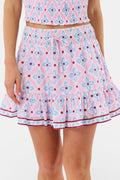 A woman wears the Roller Rabbit Lerato Peyton Skirt, a pink cotton skirt with a blue and red geometric floral pattern, ruffled hem, and matching sleeveless crop top for a summery, casual look.