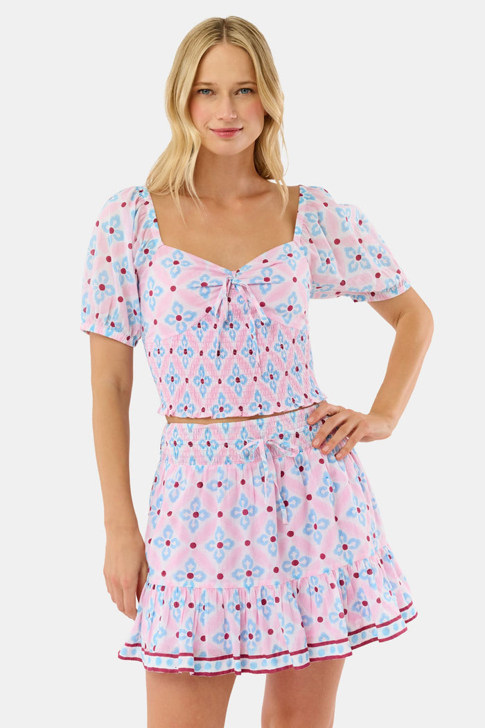 A woman wears the Roller Rabbit Lerato Peyton Skirt, a pink cotton skirt with a blue and red geometric floral pattern, ruffled hem, and matching sleeveless crop top for a summery, casual look.