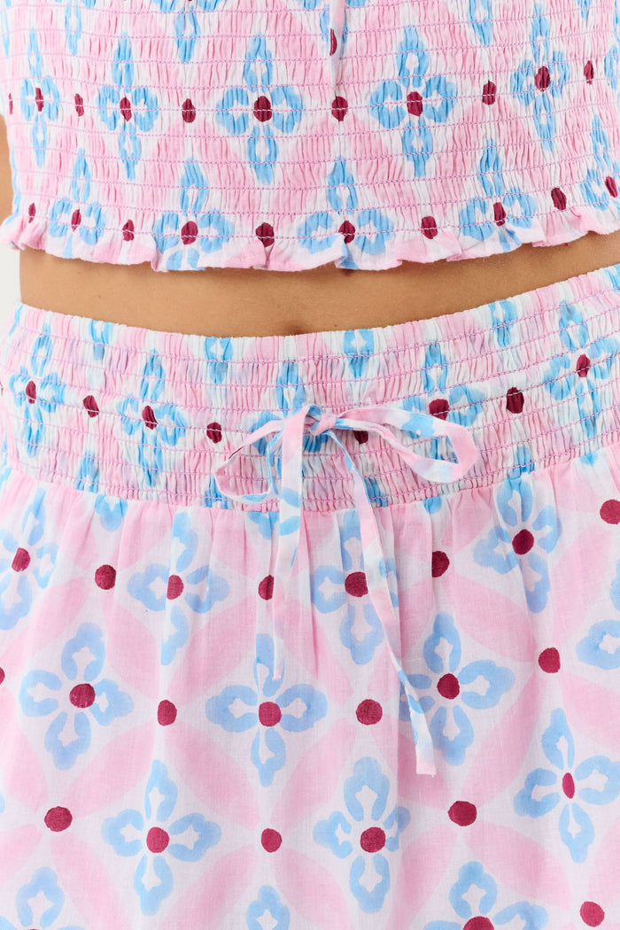 A woman wears the Roller Rabbit Lerato Peyton Skirt, a pink cotton skirt with a blue and red geometric floral pattern, ruffled hem, and matching sleeveless crop top for a summery, casual look.