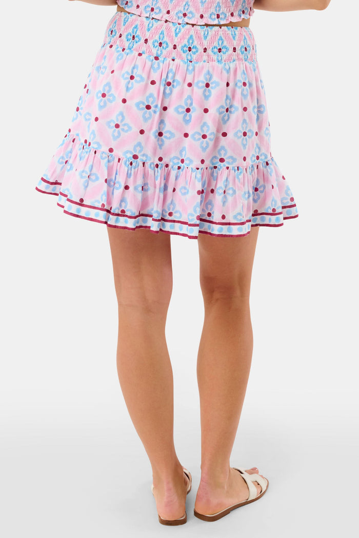 A woman wears the Roller Rabbit Lerato Peyton Skirt, a pink cotton skirt with a blue and red geometric floral pattern, ruffled hem, and matching sleeveless crop top for a summery, casual look.
