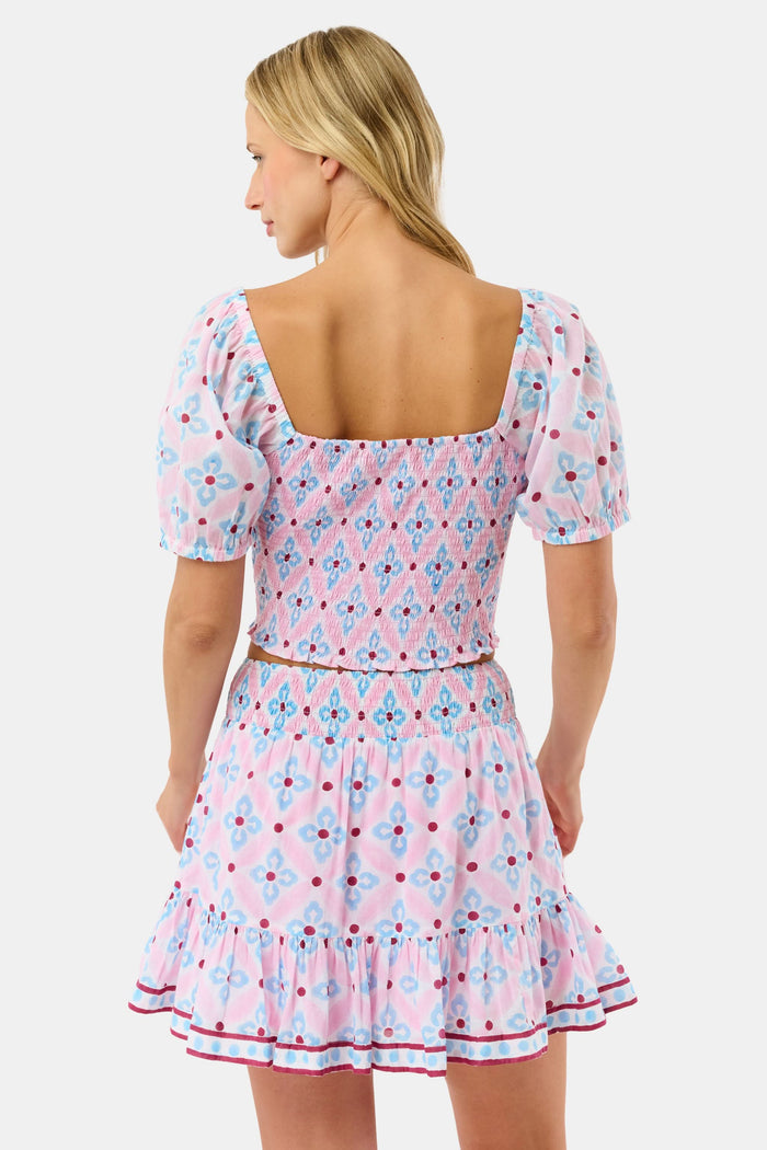 A woman wears the Roller Rabbit Lerato Peyton Skirt, a pink cotton skirt with a blue and red geometric floral pattern, ruffled hem, and matching sleeveless crop top for a summery, casual look.