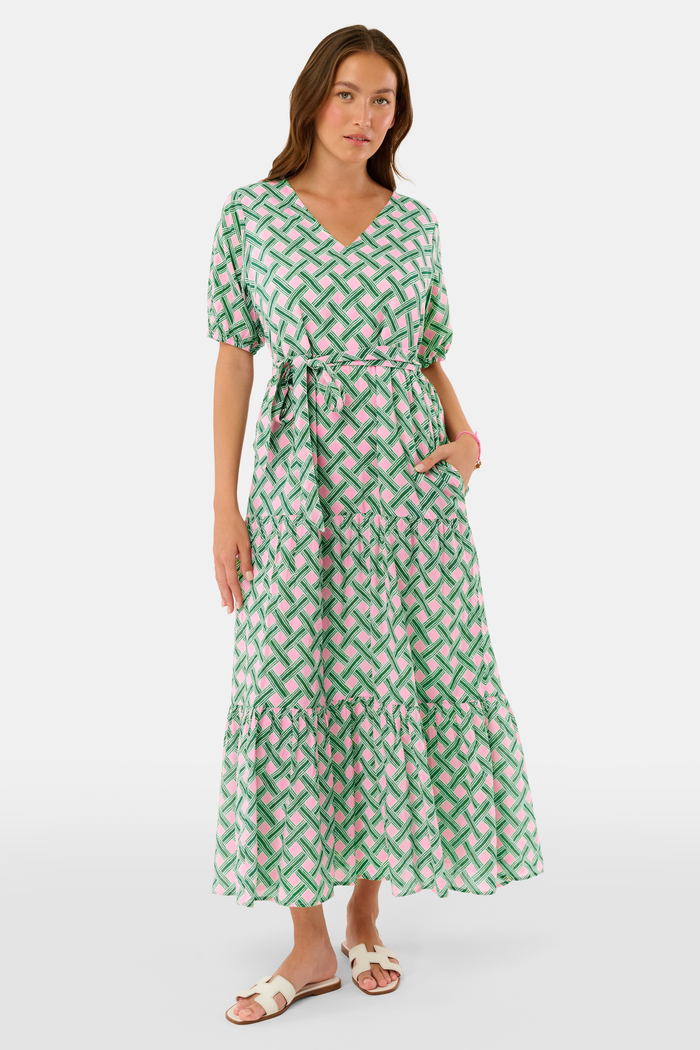 A woman models the Roller Rabbit Liana Brigitta Dress, a cotton voile maxi in pink and green Liana print, featuring short sleeves, a V-neck, and tie waist. She pairs it with white sandals and has wavy brown hair.