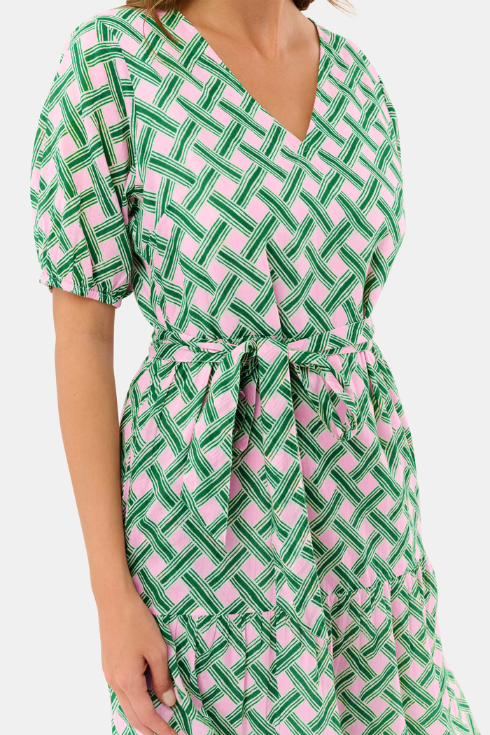 A woman models the Roller Rabbit Liana Brigitta Dress, a cotton voile maxi in pink and green Liana print, featuring short sleeves, a V-neck, and tie waist. She pairs it with white sandals and has wavy brown hair.