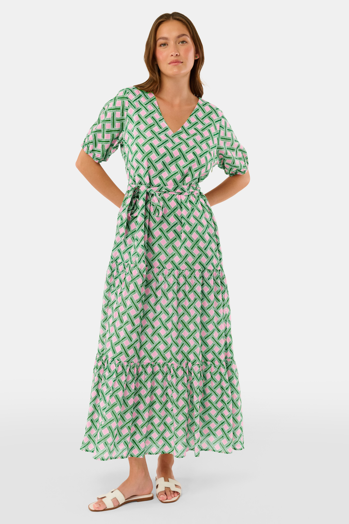 A woman models the Roller Rabbit Liana Brigitta Dress, a cotton voile maxi in pink and green Liana print, featuring short sleeves, a V-neck, and tie waist. She pairs it with white sandals and has wavy brown hair.