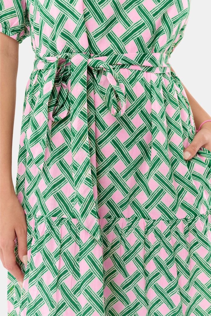 A woman models the Roller Rabbit Liana Brigitta Dress, a cotton voile maxi in pink and green Liana print, featuring short sleeves, a V-neck, and tie waist. She pairs it with white sandals and has wavy brown hair.