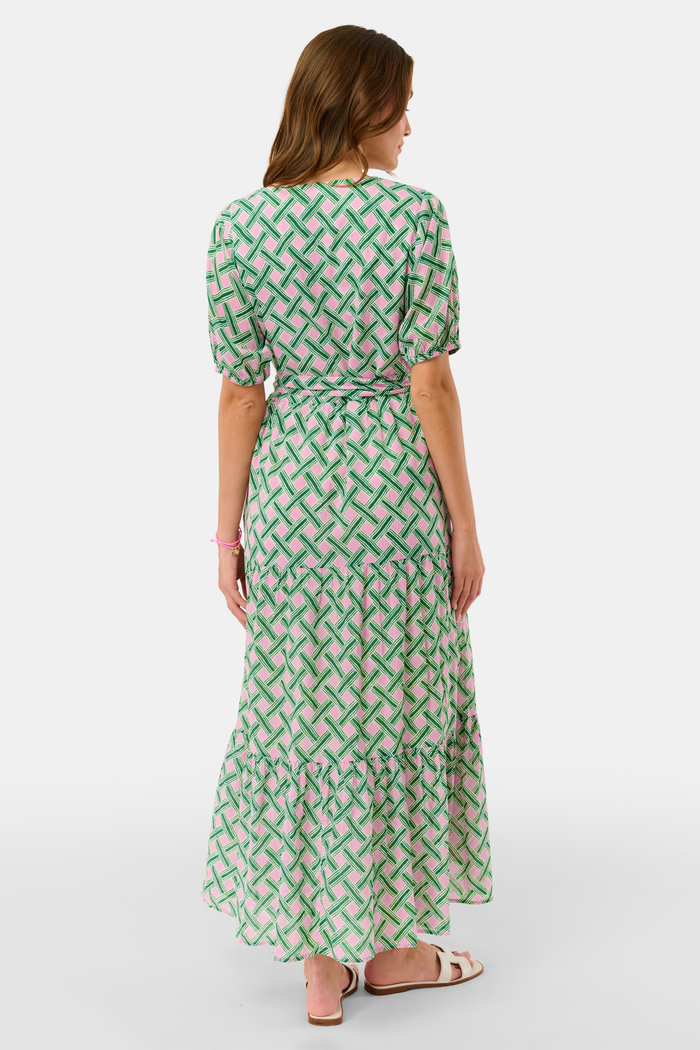 A woman models the Roller Rabbit Liana Brigitta Dress, a cotton voile maxi in pink and green Liana print, featuring short sleeves, a V-neck, and tie waist. She pairs it with white sandals and has wavy brown hair.