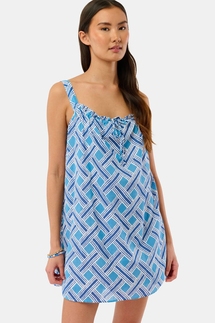 A woman models the Roller Rabbit Liana Keaton Dress, a blue and white cotton sundress with a geometric pattern and ruffled neckline, standing against a plain light background.