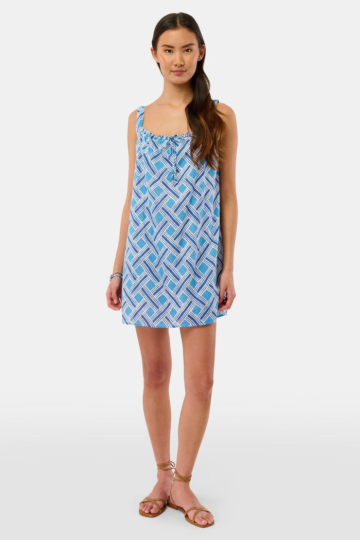 A woman models the Roller Rabbit Liana Keaton Dress, a blue and white cotton sundress with a geometric pattern and ruffled neckline, standing against a plain light background.