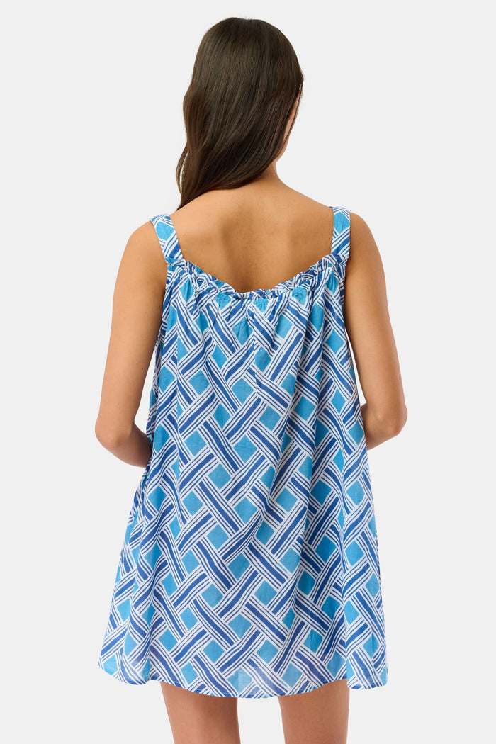 A woman models the Roller Rabbit Liana Keaton Dress, a blue and white cotton sundress with a geometric pattern and ruffled neckline, standing against a plain light background.