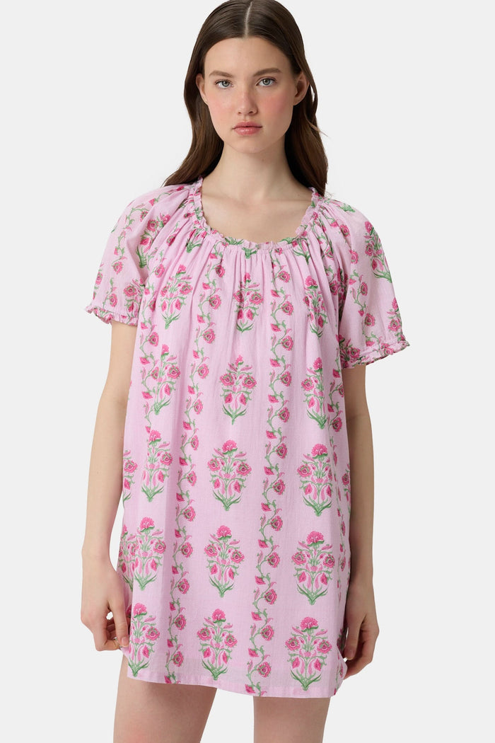 A woman with long brown hair wears the Roller Rabbit Lily Stripe Briella Dress, a loose cotton voile lounge dress with puffed short sleeves and a green and pink floral pattern, standing against a plain light background.