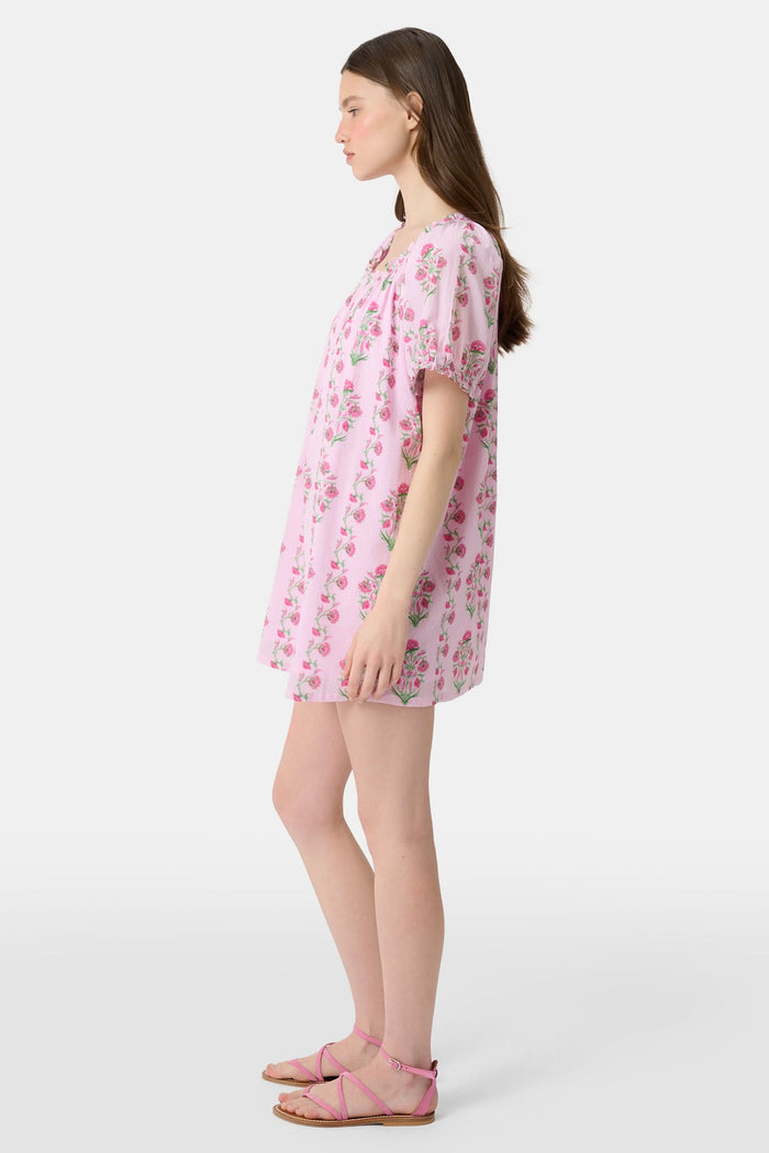 A woman with long brown hair wears the Roller Rabbit Lily Stripe Briella Dress, a loose cotton voile lounge dress with puffed short sleeves and a green and pink floral pattern, standing against a plain light background.