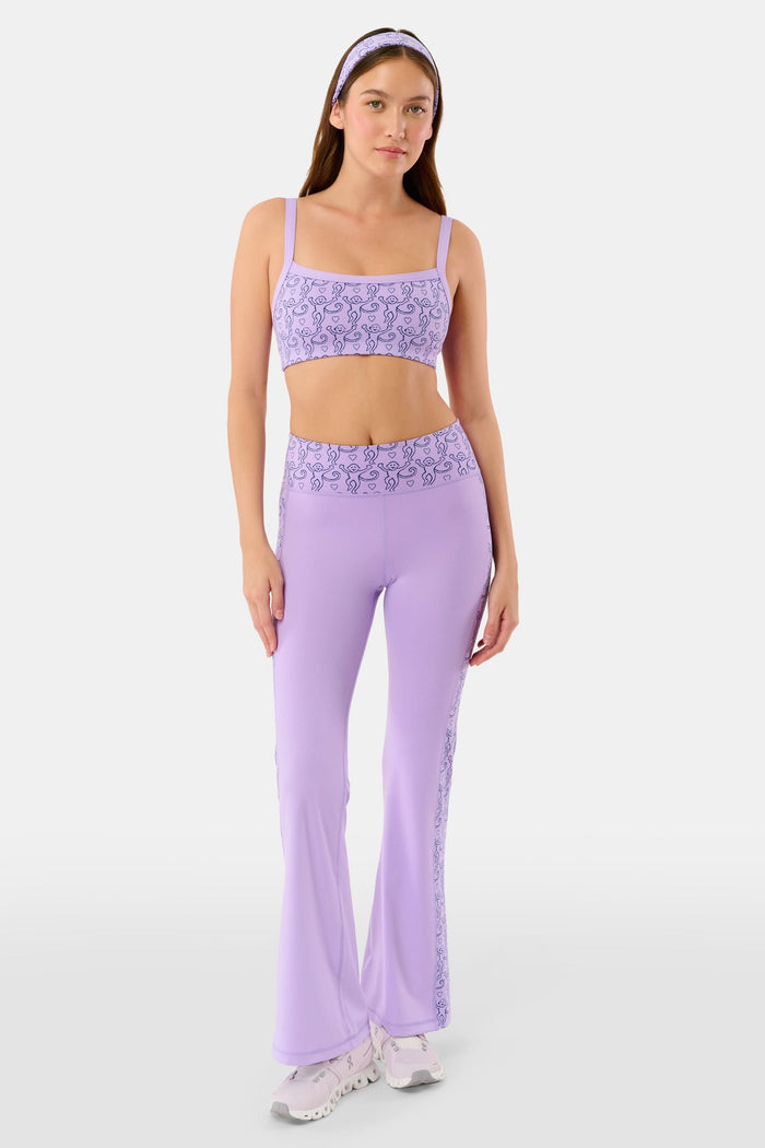 A person wears Roller Rabbit’s Lovely Monkey Ally Flared Leggings—lavender with a patterned waistband and side panels—paired with white sneakers, standing against a plain white background.