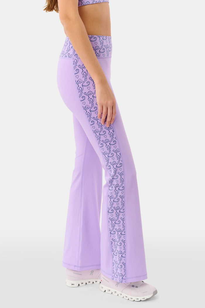 A person wears Roller Rabbit’s Lovely Monkey Ally Flared Leggings—lavender with a patterned waistband and side panels—paired with white sneakers, standing against a plain white background.