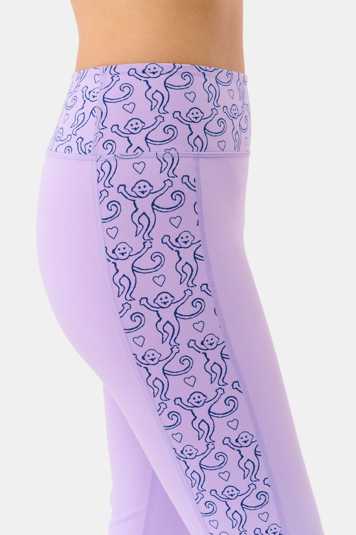 A person wears Roller Rabbit’s Lovely Monkey Ally Flared Leggings—lavender with a patterned waistband and side panels—paired with white sneakers, standing against a plain white background.