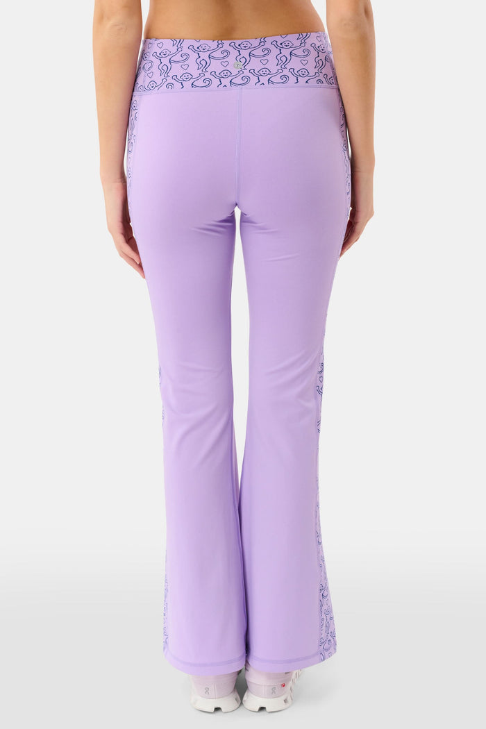 A person wears Roller Rabbit’s Lovely Monkey Ally Flared Leggings—lavender with a patterned waistband and side panels—paired with white sneakers, standing against a plain white background.