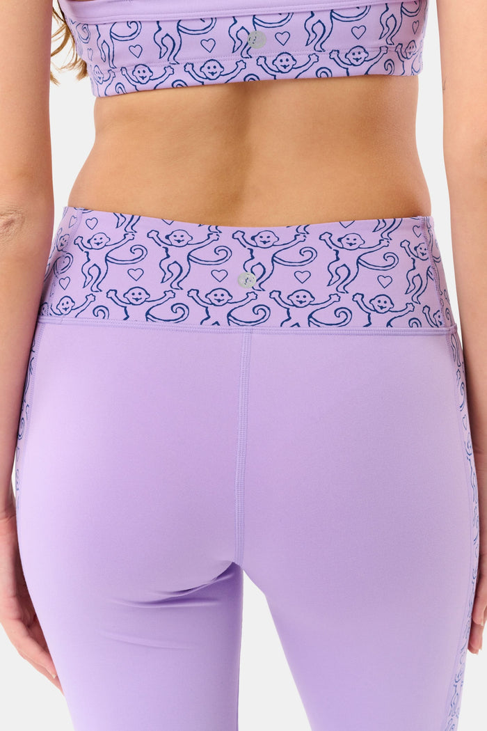 A person wears Roller Rabbit’s Lovely Monkey Ally Flared Leggings—lavender with a patterned waistband and side panels—paired with white sneakers, standing against a plain white background.