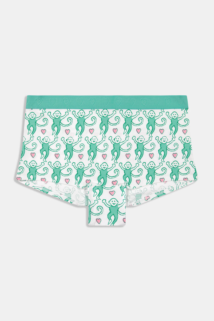 A woman with long brown hair wears Roller Rabbit’s Lovely Monkey Crosby Boyshort and matching sports bra with a white base, green trim, and monkey print, standing against a plain white background.