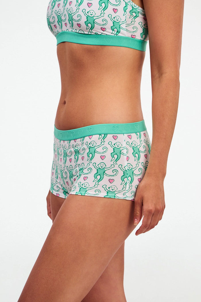 A woman with long brown hair wears Roller Rabbit’s Lovely Monkey Crosby Boyshort and matching sports bra with a white base, green trim, and monkey print, standing against a plain white background.