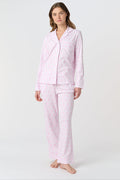 A woman with light brown hair and a neutral expression stands barefoot on a white background wearing Roller Rabbit's Lovely Monkey Long Sleeve Polo Pajamas, a light pink and white 100 Pima Cotton set.