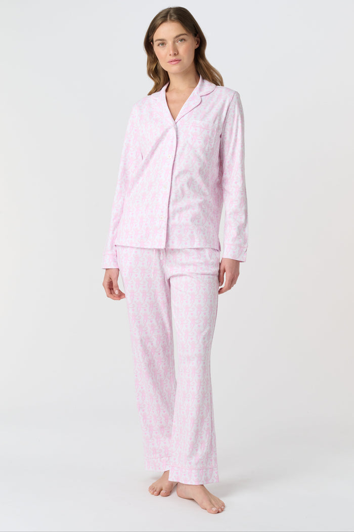 A woman with light brown hair and a neutral expression stands barefoot on a white background wearing Roller Rabbit's Lovely Monkey Long Sleeve Polo Pajamas, a light pink and white 100 Pima Cotton set.