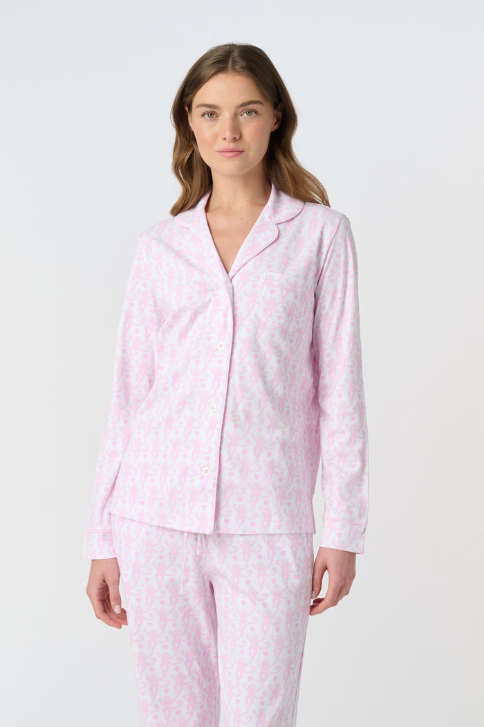 A woman with light brown hair and a neutral expression stands barefoot on a white background wearing Roller Rabbit's Lovely Monkey Long Sleeve Polo Pajamas, a light pink and white 100 Pima Cotton set.