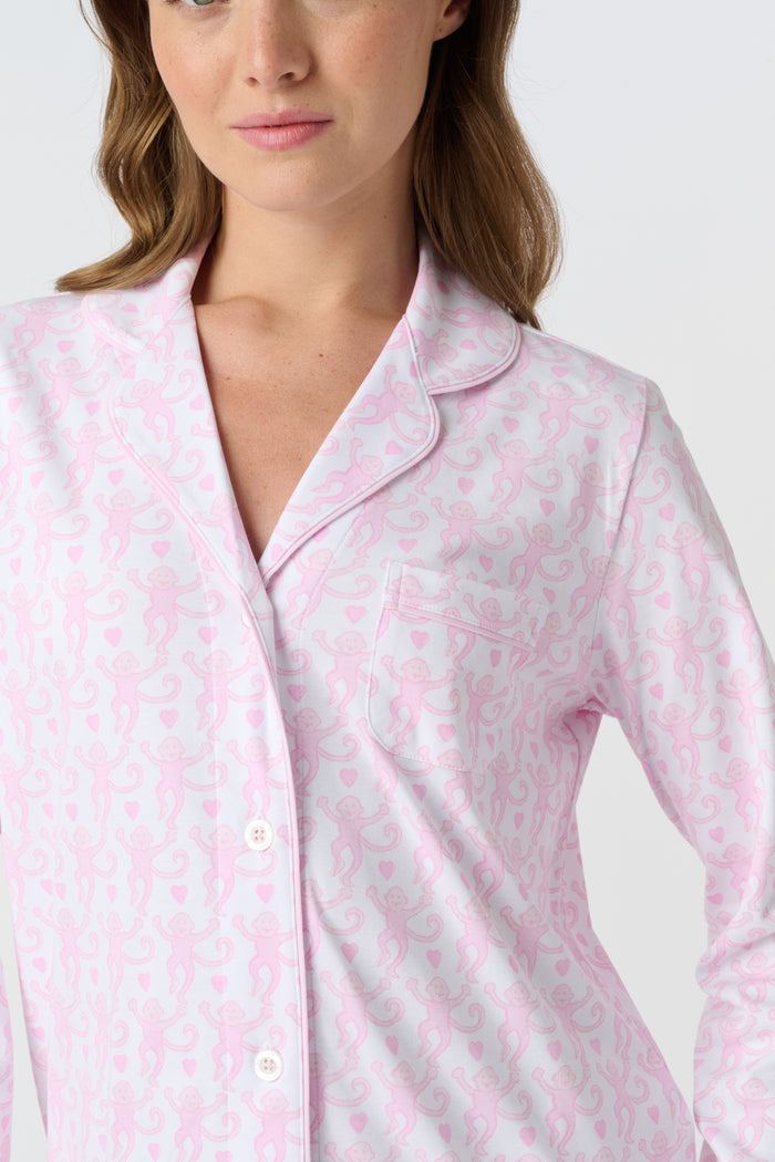 A woman with light brown hair and a neutral expression stands barefoot on a white background wearing Roller Rabbit's Lovely Monkey Long Sleeve Polo Pajamas, a light pink and white 100 Pima Cotton set.