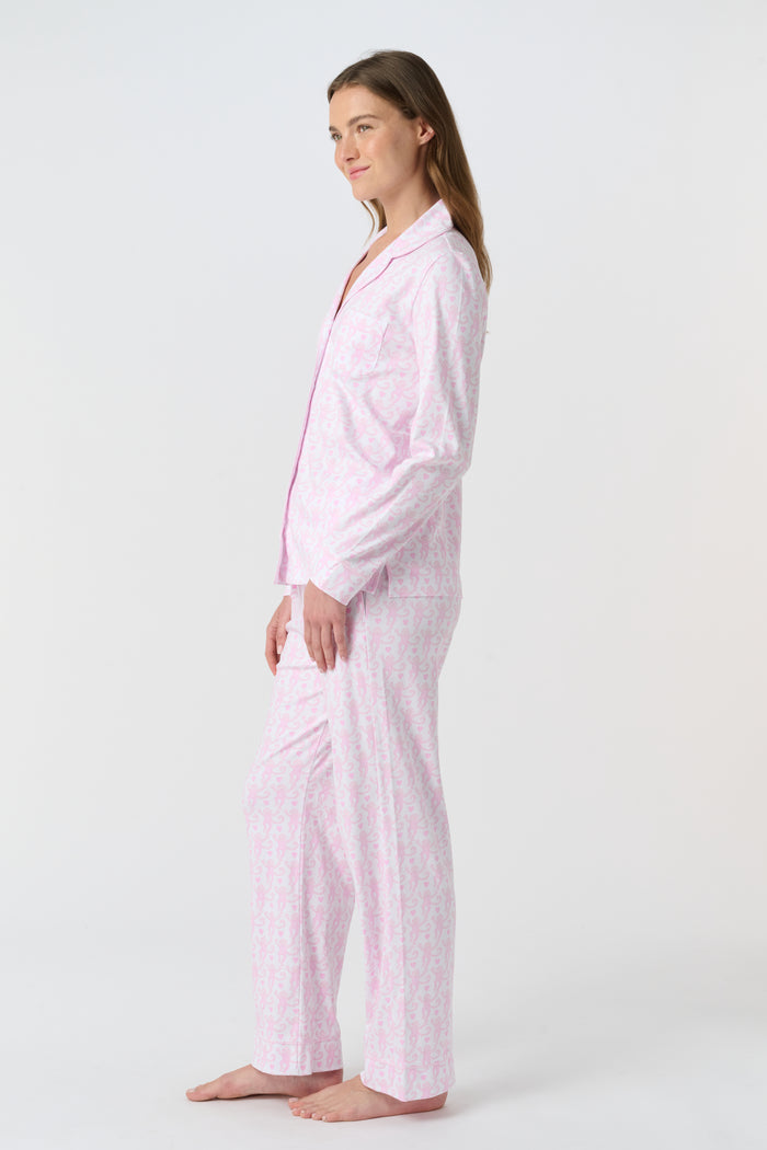 A woman with light brown hair and a neutral expression stands barefoot on a white background wearing Roller Rabbit's Lovely Monkey Long Sleeve Polo Pajamas, a light pink and white 100 Pima Cotton set.