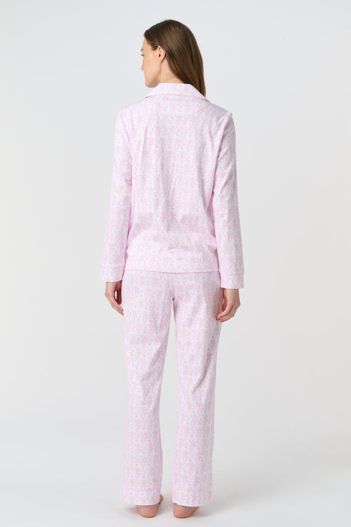 A woman with light brown hair and a neutral expression stands barefoot on a white background wearing Roller Rabbit's Lovely Monkey Long Sleeve Polo Pajamas, a light pink and white 100 Pima Cotton set.