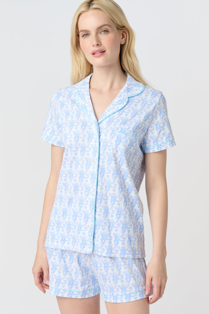 A woman with long blonde hair wears the Roller Rabbit Lovely Monkey Polo Pajamas set in light blue and white Pima cotton shorts, smiling softly against a plain light background.