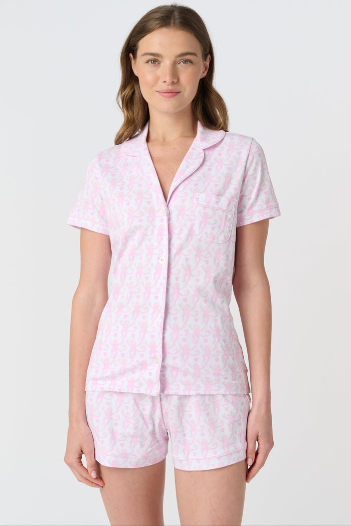 A woman wearing the Roller Rabbit Lovely Monkey Polo Pajamas—a light pink and white patterned short sleeve button-up top with matching shorts—stands against a plain white background, smiling softly.