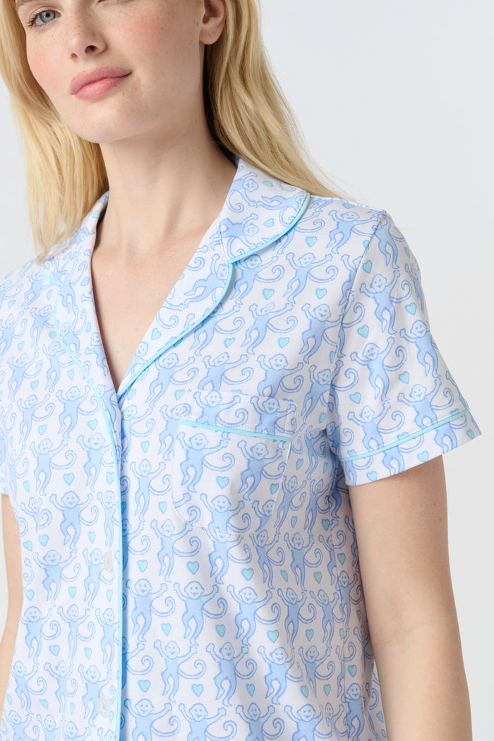 A woman with long blonde hair wears the Roller Rabbit Lovely Monkey Polo Pajamas set in light blue and white Pima cotton shorts, smiling softly against a plain light background.