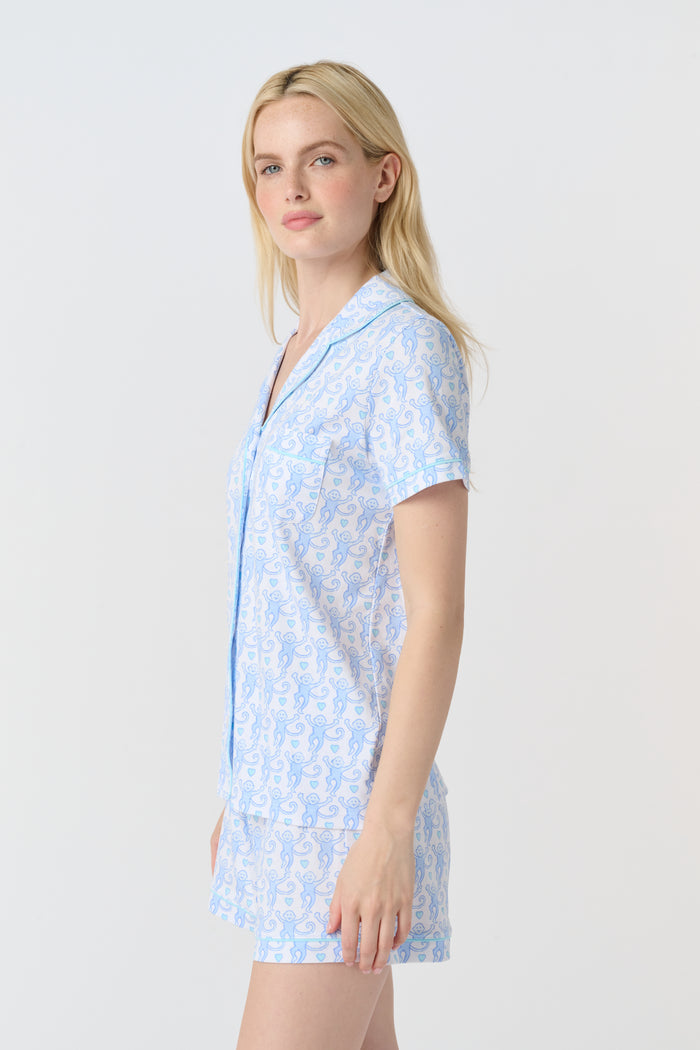 A woman with long blonde hair wears the Roller Rabbit Lovely Monkey Polo Pajamas set in light blue and white Pima cotton shorts, smiling softly against a plain light background.