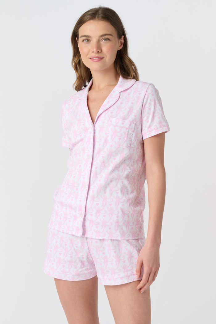 A woman wearing the Roller Rabbit Lovely Monkey Polo Pajamas—a light pink and white patterned short sleeve button-up top with matching shorts—stands against a plain white background, smiling softly.