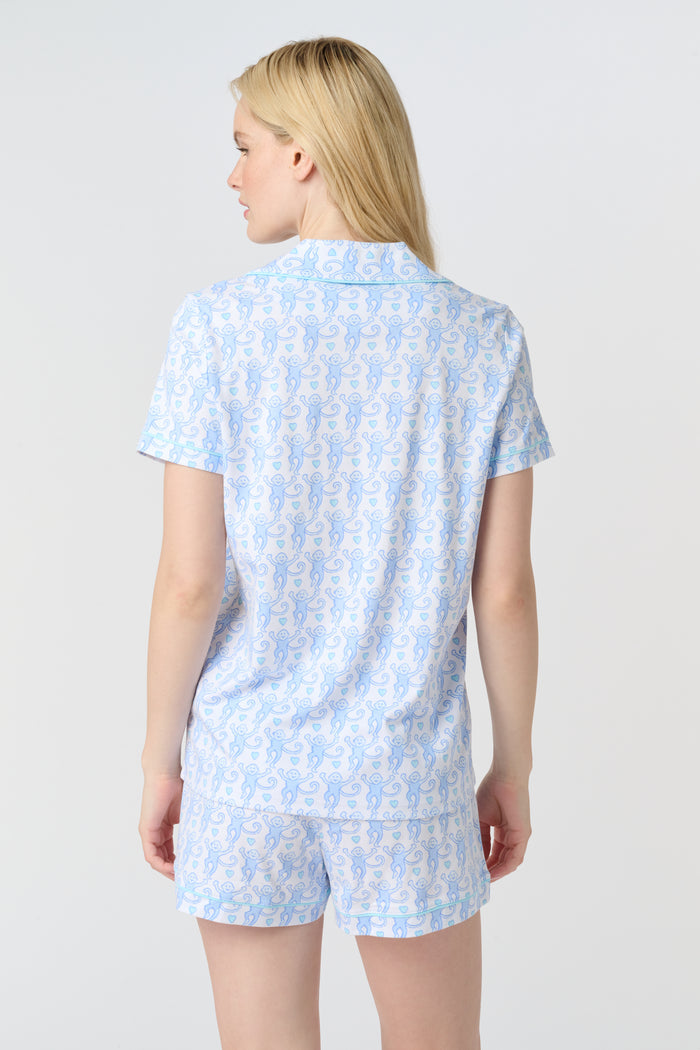 A woman with long blonde hair wears the Roller Rabbit Lovely Monkey Polo Pajamas set in light blue and white Pima cotton shorts, smiling softly against a plain light background.