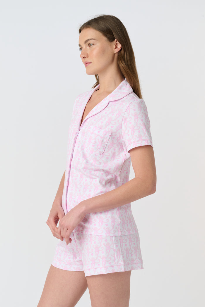 A woman wearing the Roller Rabbit Lovely Monkey Polo Pajamas—a light pink and white patterned short sleeve button-up top with matching shorts—stands against a plain white background, smiling softly.