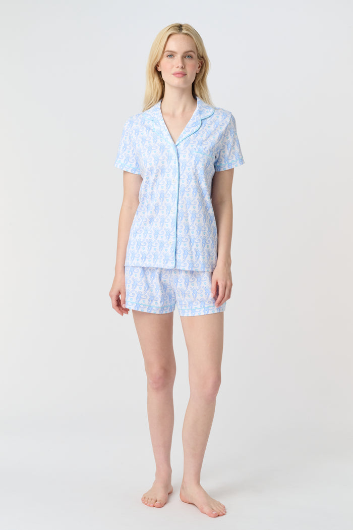 A woman with long blonde hair wears the Roller Rabbit Lovely Monkey Polo Pajamas set in light blue and white Pima cotton shorts, smiling softly against a plain light background.