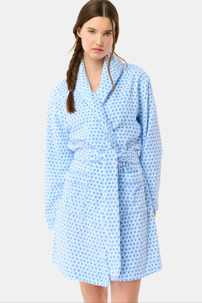A woman with a side braid wears the Roller Rabbit Lovely Stripe Max Robe, a light blue cotton piece with small blue heart patterns, belt, and large front pockets. She stands against a plain background, looking at the camera neutrally.