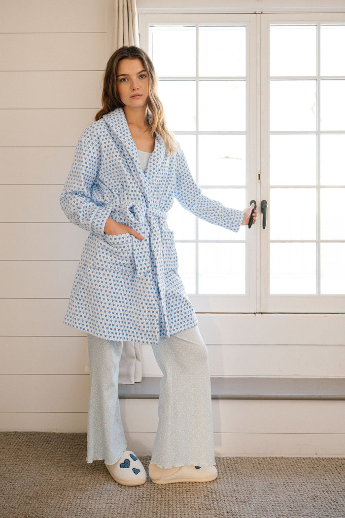 A woman with a side braid wears the Roller Rabbit Lovely Stripe Max Robe, a light blue cotton piece with small blue heart patterns, belt, and large front pockets. She stands against a plain background, looking at the camera neutrally.