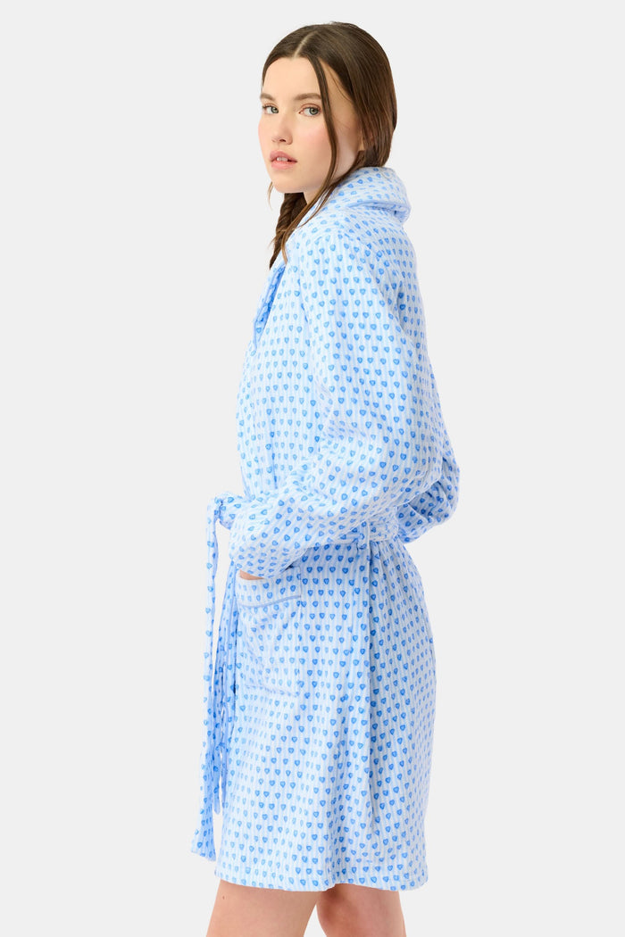 A woman with a side braid wears the Roller Rabbit Lovely Stripe Max Robe, a light blue cotton piece with small blue heart patterns, belt, and large front pockets. She stands against a plain background, looking at the camera neutrally.