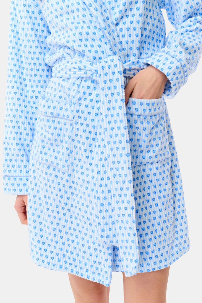 A woman with a side braid wears the Roller Rabbit Lovely Stripe Max Robe, a light blue cotton piece with small blue heart patterns, belt, and large front pockets. She stands against a plain background, looking at the camera neutrally.