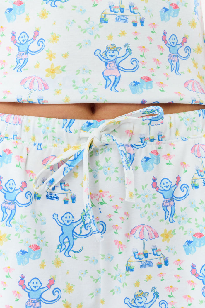 A blonde woman wears the Roller Rabbit May Flowers Mischa Set, a soft Pima cotton pajama with blue cartoon prints, pink trim, and cheerful patterns. The sleeveless top and loose shorts provide cozy comfort.