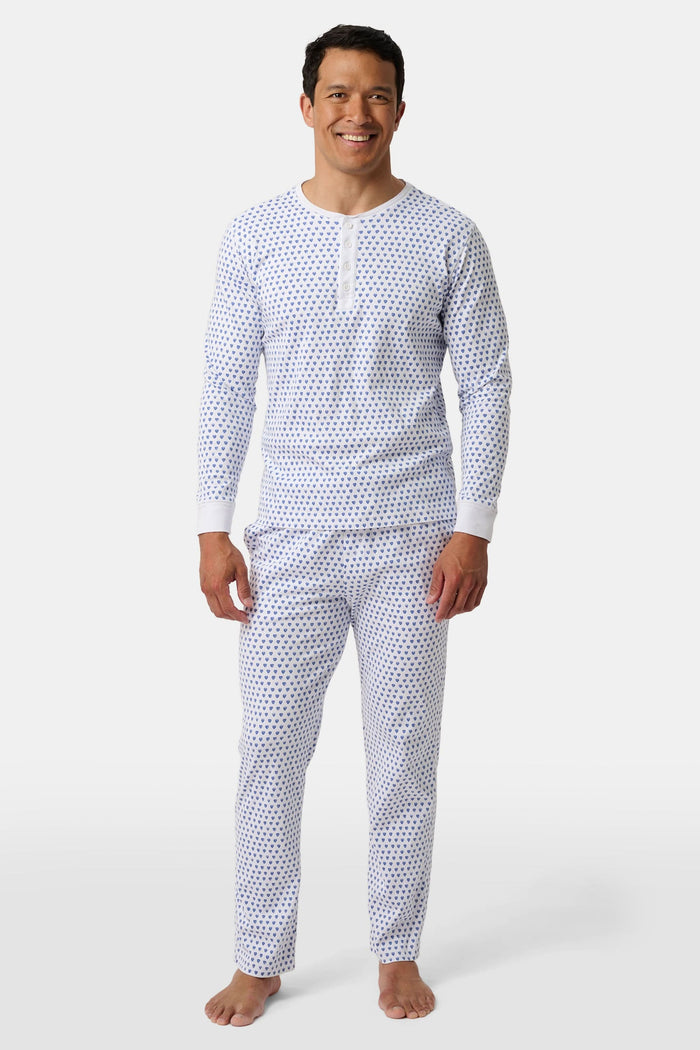 A man stands barefoot, smiling in Roller Rabbit’s Mens Hearts Spencer Pajamas. This matching set has a white background with small blue and red heart patterns. Made of 100 Pima cotton, the pajama top features comfy henley-style buttons.