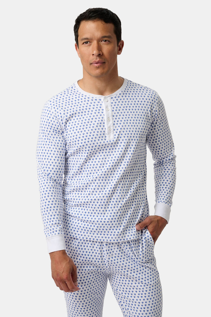 A man stands barefoot, smiling in Roller Rabbit’s Mens Hearts Spencer Pajamas. This matching set has a white background with small blue and red heart patterns. Made of 100 Pima cotton, the pajama top features comfy henley-style buttons.