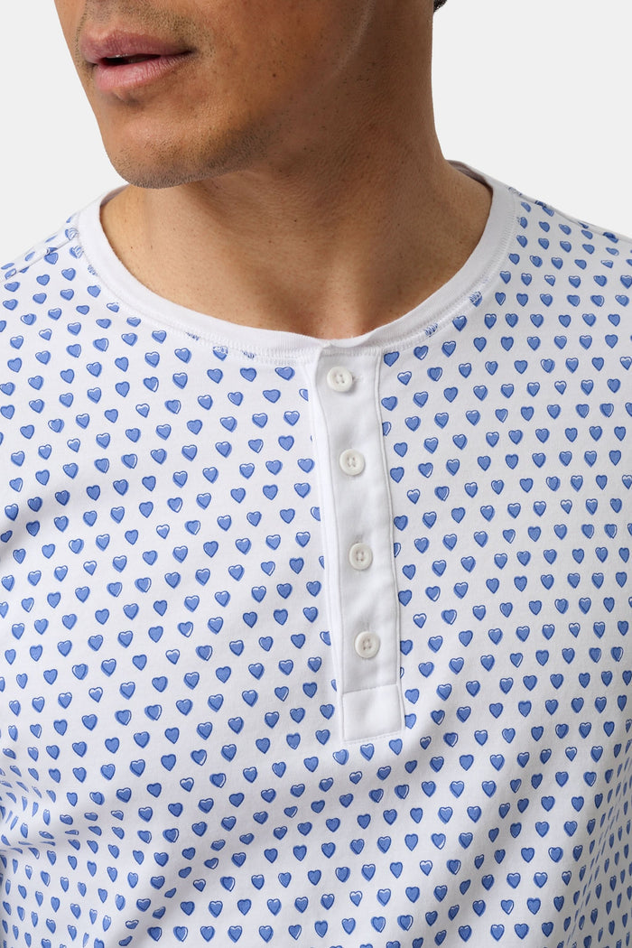 A man stands barefoot, smiling in Roller Rabbit’s Mens Hearts Spencer Pajamas. This matching set has a white background with small blue and red heart patterns. Made of 100 Pima cotton, the pajama top features comfy henley-style buttons.