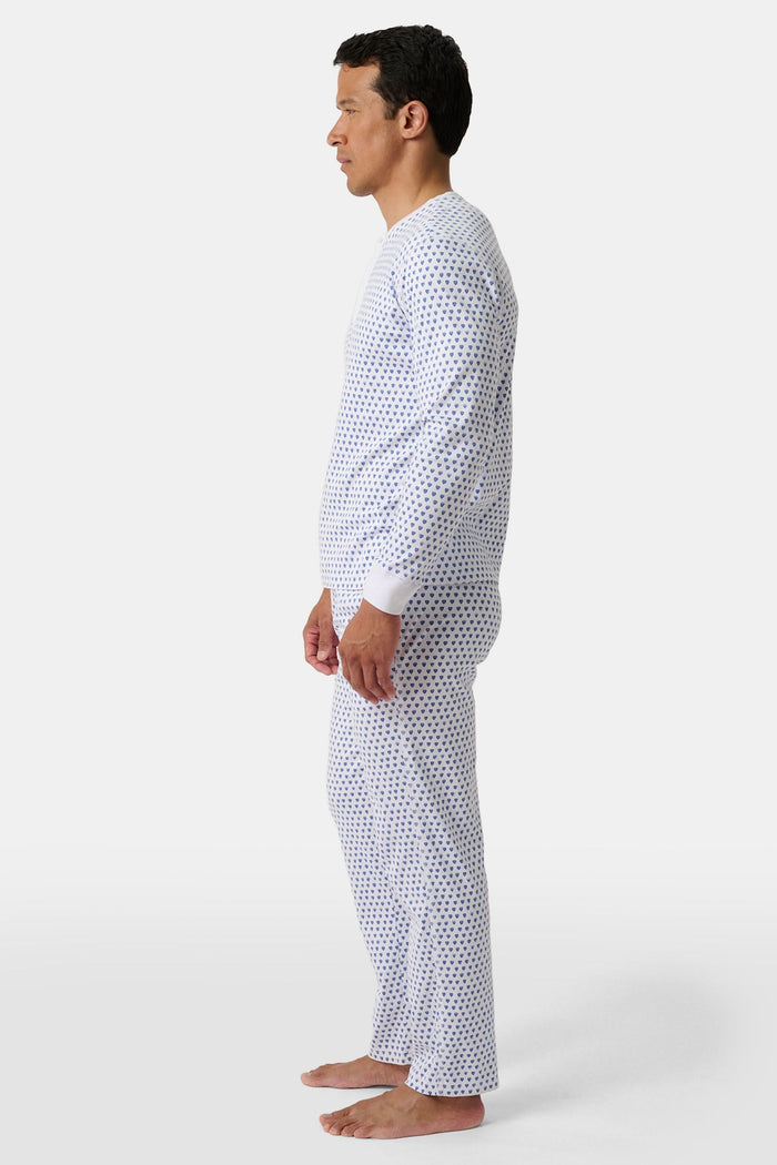 A man stands barefoot, smiling in Roller Rabbit’s Mens Hearts Spencer Pajamas. This matching set has a white background with small blue and red heart patterns. Made of 100 Pima cotton, the pajama top features comfy henley-style buttons.