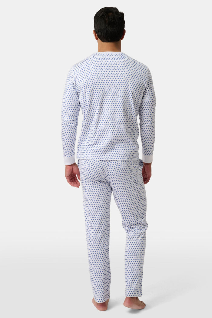 A man stands barefoot, smiling in Roller Rabbit’s Mens Hearts Spencer Pajamas. This matching set has a white background with small blue and red heart patterns. Made of 100 Pima cotton, the pajama top features comfy henley-style buttons.