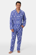 A man stands barefoot in Roller Rabbit’s Mens Lobster Trellis Loungewear, a matching blue and white floral cotton voile pajama set with a button-up long-sleeve shirt and pants. He has one hand in his pocket and looks forward.